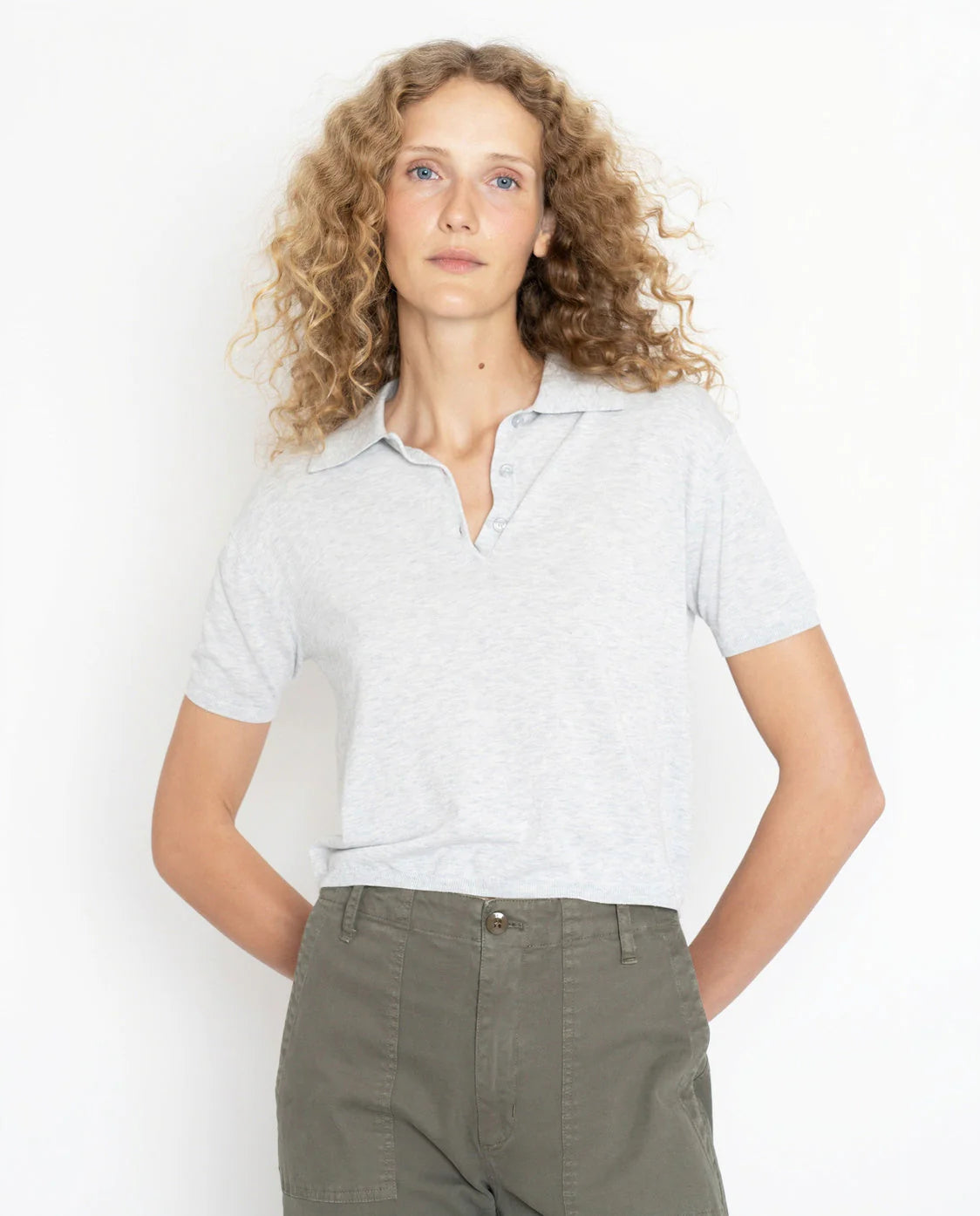 A woman with blonde curly hair wears The Farley, a light gray short-sleeve polo sweater by Kule, crafted from a lightweight cotton and silk blend, tucked into high-waisted olive green pants against a white background.