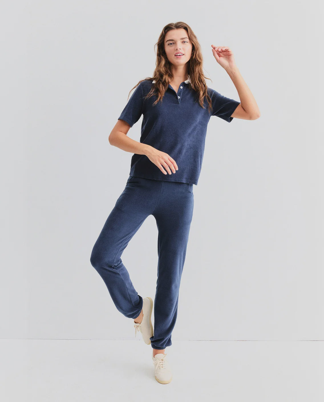 A woman models Kule's The Terry Sweatpants, paired with a navy short-sleeve top, posing on one leg and smiling against a plain white background.