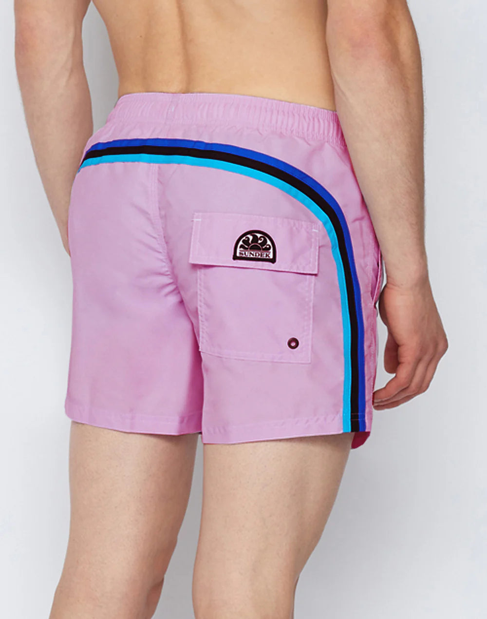 Someone shown from behind wears SUNDEK/KickoffUSA ELASTIC WAIST Board Shorts in light pink, featuring blue and dark blue side stripes and a back flap pocket, set against a plain background.