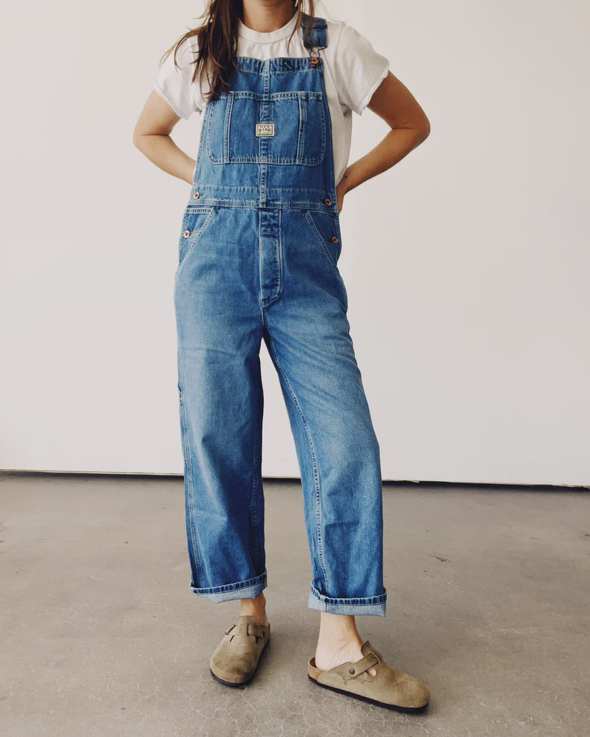 Wearing a white t-shirt and "The Miner Overalls" from Hey Gang, featuring a vintage washed denim look with an oversized fit, the individual stands confidently with hands on hips. Complemented by brown slip-on shoes, this unisex ensemble is set against a plain, light-colored wall and concrete floor.