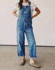 Wearing a white t-shirt and "The Miner Overalls" from Hey Gang, featuring a vintage washed denim look with an oversized fit, the individual stands confidently with hands on hips. Complemented by brown slip-on shoes, this unisex ensemble is set against a plain, light-colored wall and concrete floor.