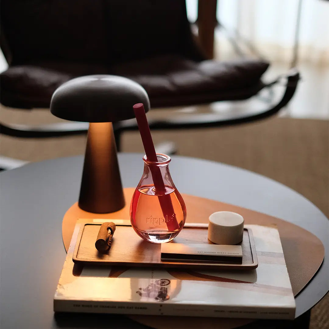 A small glass with a red drink and straw sits on a wooden tray beside the Faire Boost Reed Diffuser, atop stacked books next to a modern black lamp on a round table, with a leather chair in the background.