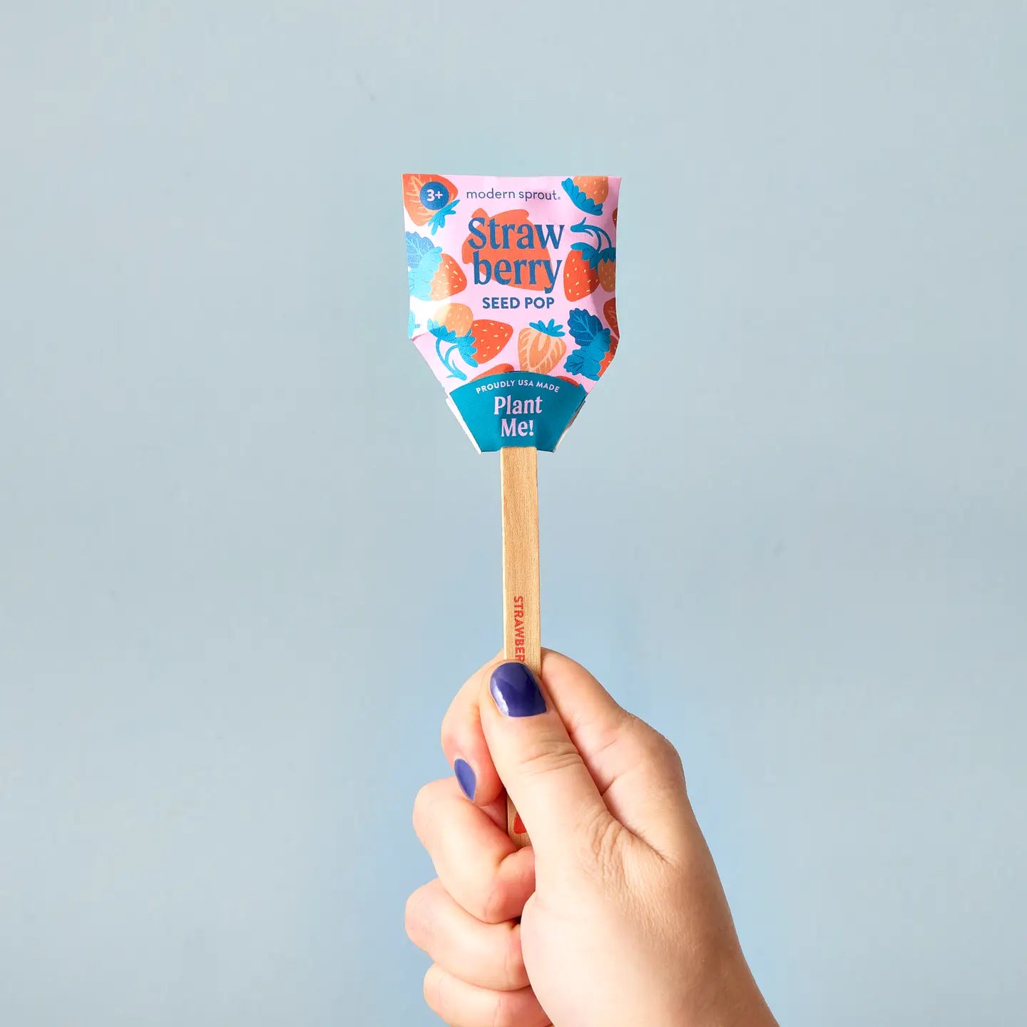 A hand with blue nail polish holds a Faire Garden Seed Pop in vibrant strawberry-themed packaging labeled “Plant Me!”—a playful way to start your vegetable garden—against a light blue background.