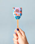 A hand with blue nail polish holds a Faire Garden Seed Pop in vibrant strawberry-themed packaging labeled “Plant Me!”—a playful way to start your vegetable garden—against a light blue background.