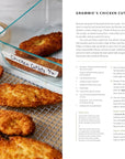 A hand lifts a breaded chicken cutlet from a baking sheet with a fork. Golden-brown cutlets cool on a rack next to a labeled Every Day With Babs glass dish by Random House—ideal for comforting family meals.