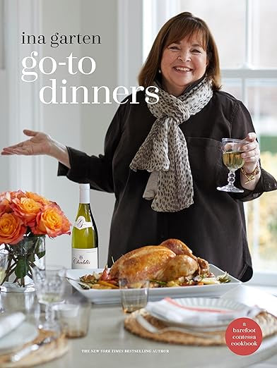 A woman beams delightfully, holding a glass of white wine next to a table adorned with roasted chicken and a bottle of wine. A vibrant bouquet of orange roses enhances the scene. The cover text declares "Ina Garten: Go-To Dinners" from Random House, an ideal choice for home cooks seeking simplicity and elegance in their meals.