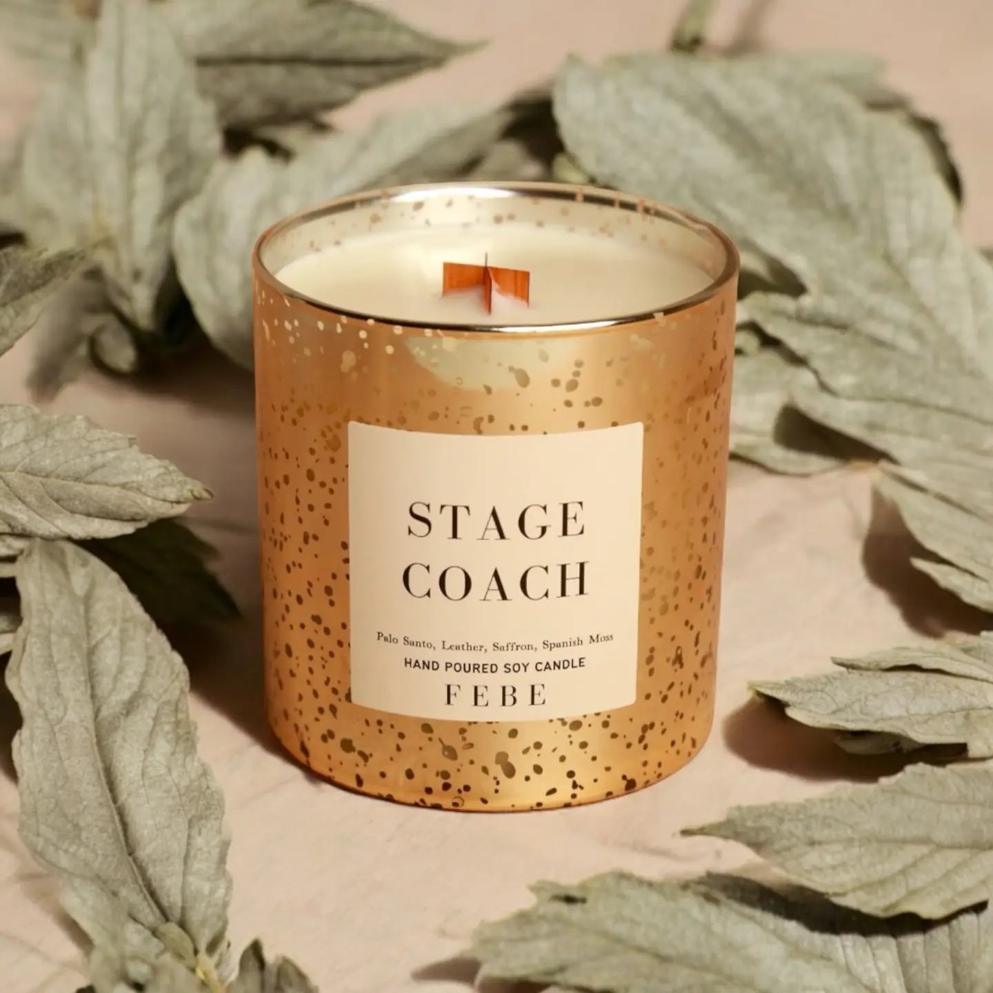 A decorative candle from the FEBE Candles collection by Faire, housed in a gold-speckled glass holder and labeled "Stage Coach," emits oud notes. The scene is complemented by dried sage leaves in the background, adding subtle hints of smoke and spice.