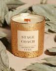 A decorative candle from the FEBE Candles collection by Faire, housed in a gold-speckled glass holder and labeled "Stage Coach," emits oud notes. The scene is complemented by dried sage leaves in the background, adding subtle hints of smoke and spice.