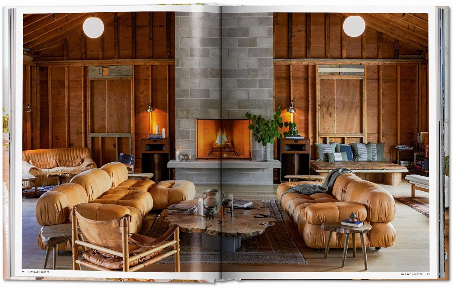 A cozy living room with tan leather sofas, a rustic wooden coffee table, and a fireplace creates the perfect setting to enjoy Great Escapes USA by Ingram Book Company. Exposed beams and shelves with plants and books add extra charm.