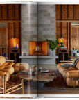 A cozy living room with tan leather sofas, a rustic wooden coffee table, and a fireplace creates the perfect setting to enjoy Great Escapes USA by Ingram Book Company. Exposed beams and shelves with plants and books add extra charm.