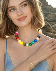 A woman with shoulder-length brown hair wears the PERRY Styles Bubble Gum Necklace, gold bracelets, and a ring. She smiles softly outdoors against rocks, dressed in a light blue spaghetti strap top.