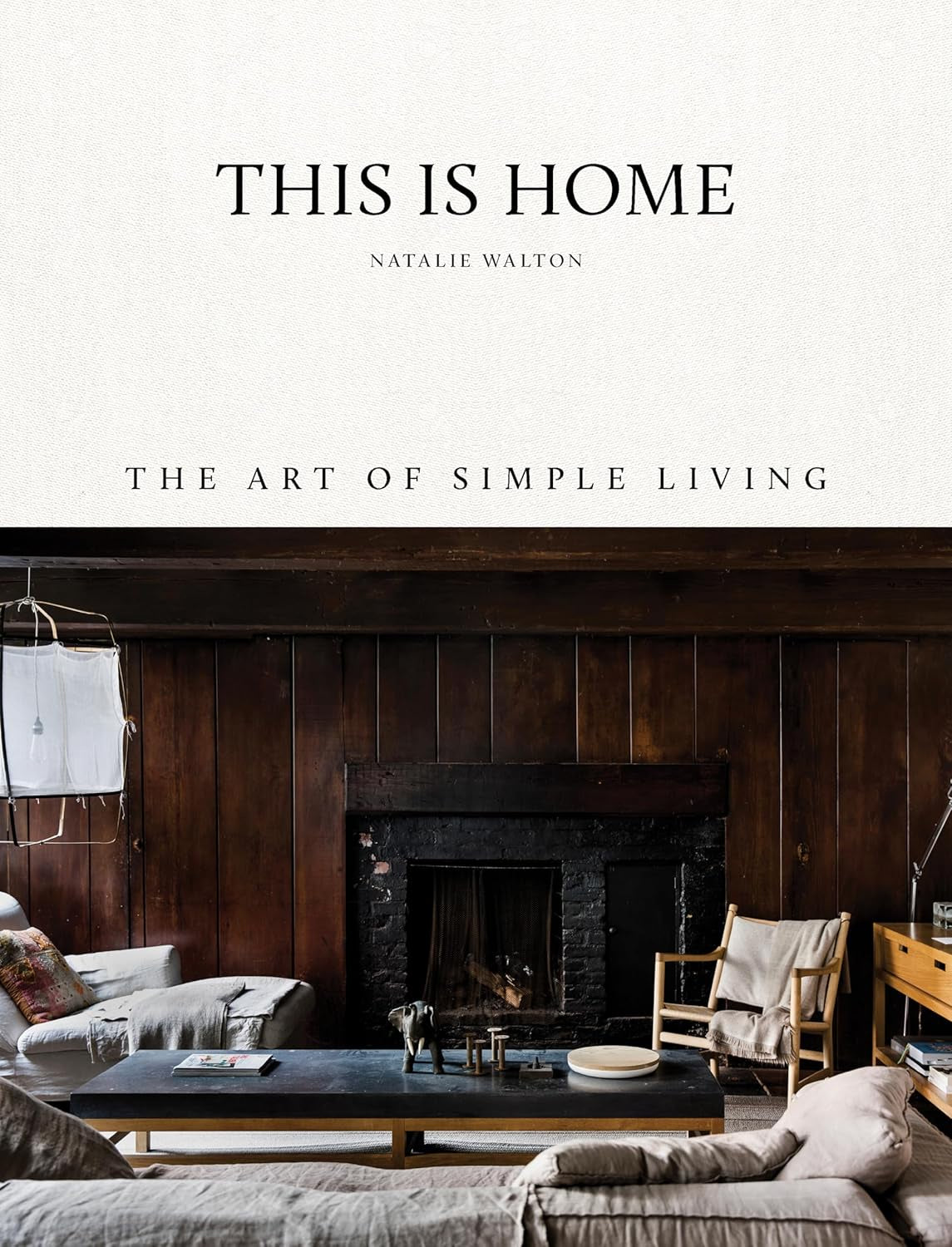 The book "This Is Home" by Random House features a cozy living room on its cover, highlighting wooden walls, a fireplace, and a cream sofa with a throw. A coffee table with books, a wooden chair, and minimal decor capture the essence of simple living.