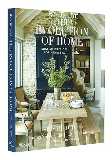 The cover of "Evolution Of Home" by Random House showcases a cozy, rustic dining area that embodies English interior design. A wooden table coupled with relaxed elegant interiors and stone walls fosters an inviting atmosphere, enhanced by books and decor that introduce a contemporary living touch.