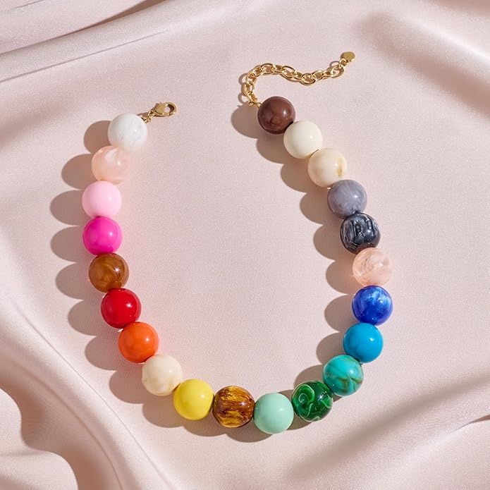 The PERRY Styles Bubble Gum Necklace is a vibrant statement piece for women, featuring large round gradient beads on a gold chain, displayed on pale pink fabric—perfect for adding a boho flair to any outfit.