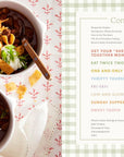 Two chili bowls with sour cream, cheese, green onions, and corn chips sit on a red-patterned cloth beside an open cookbook—perfect family meal inspiration from Every Day With Babs by Random House.