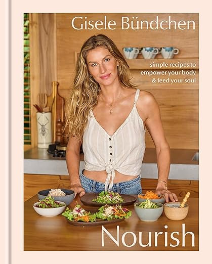 A woman with long hair stands in a kitchen, smiling over an array of wellness recipes. She wears a sleeveless top and jeans. The book cover reads "Nourish" by Random House, offering simple recipes to empower body and soul, ideal for embracing a healthy lifestyle.