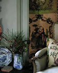 A Year in the French Style" by Random House rests on a dark table beside an upholstered armchair with floral and plaid pillows, set against a patterned wall adorned with artisanal accents and a vintage tapestry.