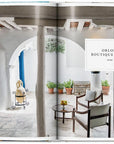 A magazine spread from "Great Escapes Greece The Hotel Book" by Ingram Book Company showcases a bright courtyard at Orloff Boutique Hotel, with potted plants, rustic beams, blue doors, and Mediterranean decor for a peaceful retreat.