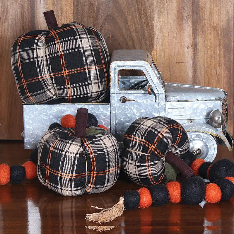 A Faire Pumpkin is displayed as farmhouse decor with a metal toy truck, three plaid pumpkins, and a black and orange felt ball garland, arranged on a wooden surface against a wooden background.