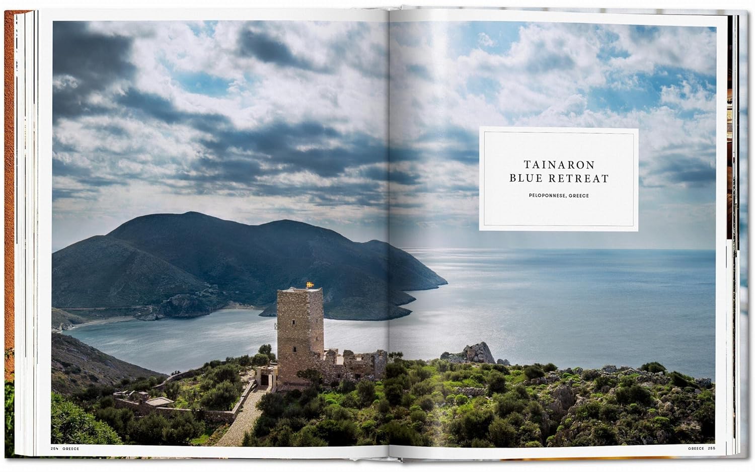 A scenic coastal view from Great Escapes Mediterranean by Ingram Book Company highlights Tainaron Blue Retreat’s stunning stone tower atop a hill overlooking the sea, mountains, and blue water beneath a partly cloudy sky.