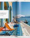 A sunlit terrace with colorful chairs and umbrellas overlooks the calm sea on Kastelorizo, as seen in "Great Escapes Greece The Hotel Book" by Ingram Book Company—featuring serene landscapes, a docked boat, and distant mountains.