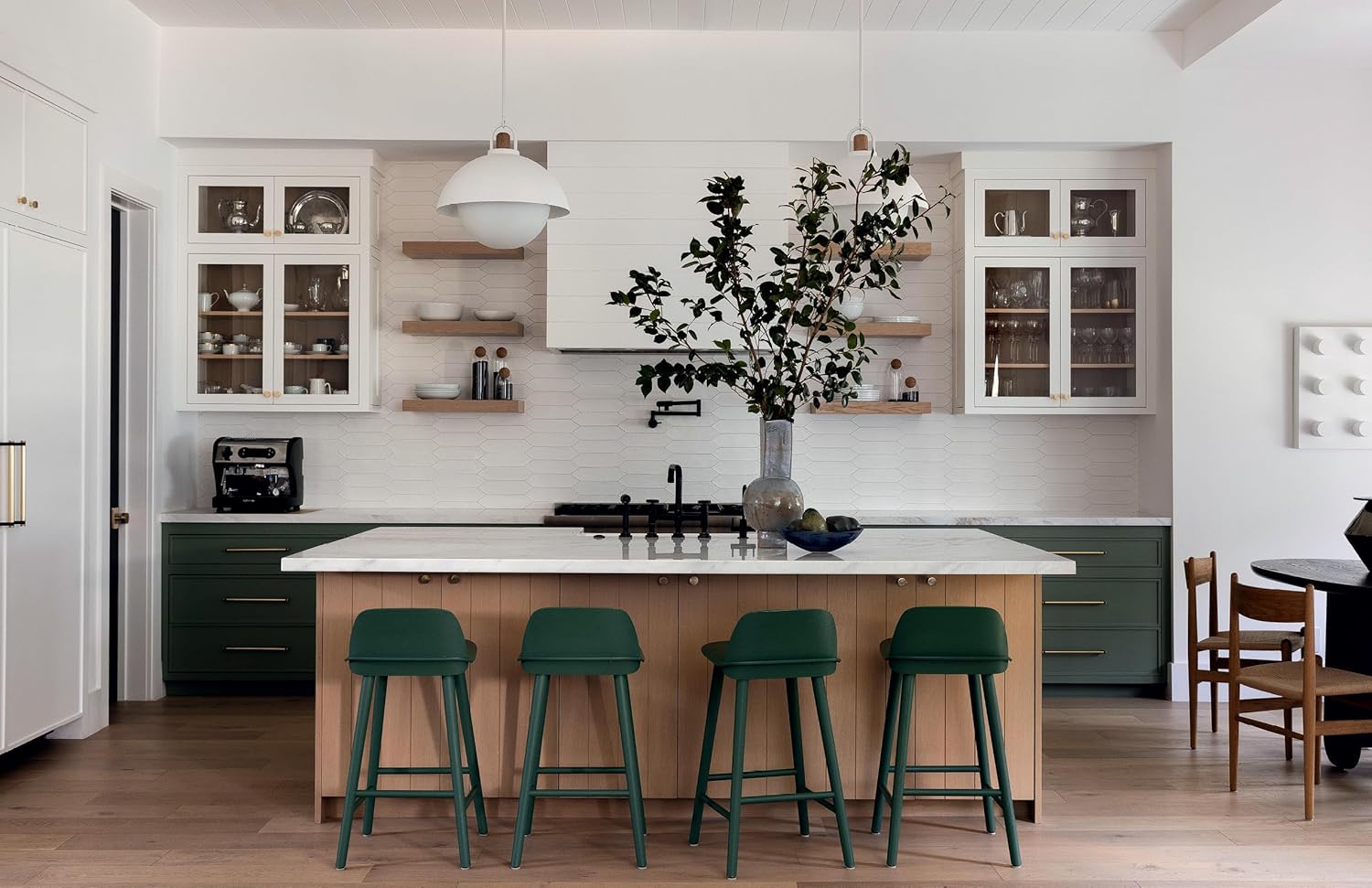 Create a nature-inspired kitchen with green cabinets, a light wood island, four green bar stools, white countertops, open shelves with dishes, pendant lights, and a large vase—style ideas from "Outside In" by Gibbs Smith Publisher.