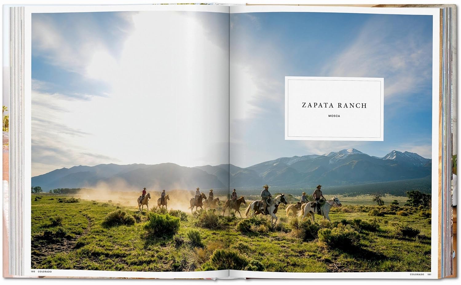 A group rides horseback across a grassy field at Zapata Ranch, one of the unique destinations showcased in Ingram Book Company’s "Great Escapes USA," with mountains and a cloudy sky stretching over two book pages.