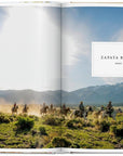 A group rides horseback across a grassy field at Zapata Ranch, one of the unique destinations showcased in Ingram Book Company’s "Great Escapes USA," with mountains and a cloudy sky stretching over two book pages.