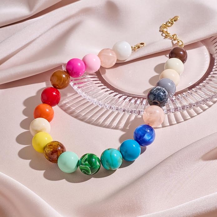 The Bubble Gum Necklace by PERRY Styles features large, round beads in a vibrant gradient, displayed on soft pink satin beside a clear textured accessory—a bold statement piece for women who love unique style.