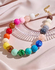 The Bubble Gum Necklace by PERRY Styles features large, round beads in a vibrant gradient, displayed on soft pink satin beside a clear textured accessory—a bold statement piece for women who love unique style.