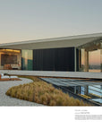 A modern glass-walled house with a cantilevered design overlooks a sunset-lit hillside, its stunning interior visible through floor-to-ceiling windows—echoing the style showcased in "Architectural Digest at 100" from Hachette Book Group.
