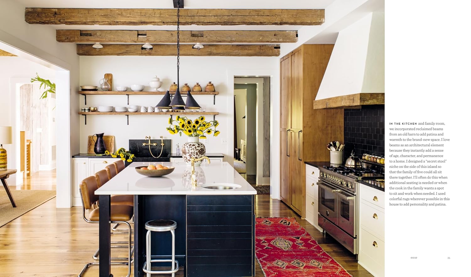 A modern kitchen featuring exposed beams, white walls, and black cabinetry channels the laid-back style of Hachette Book Group’s Down to Earth, with a large island, bar stools, pendant lights, sunflowers, and a red rug for warmth.