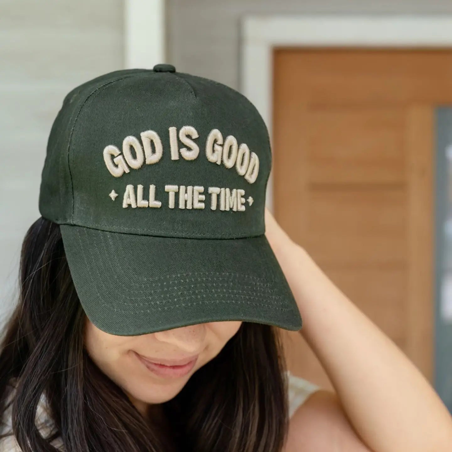 A woman with long dark hair smiles while wearing the Faire God is Good Hat, featuring green fabric and bold beige "GOD IS GOOD ALL THE TIME" embroidery.