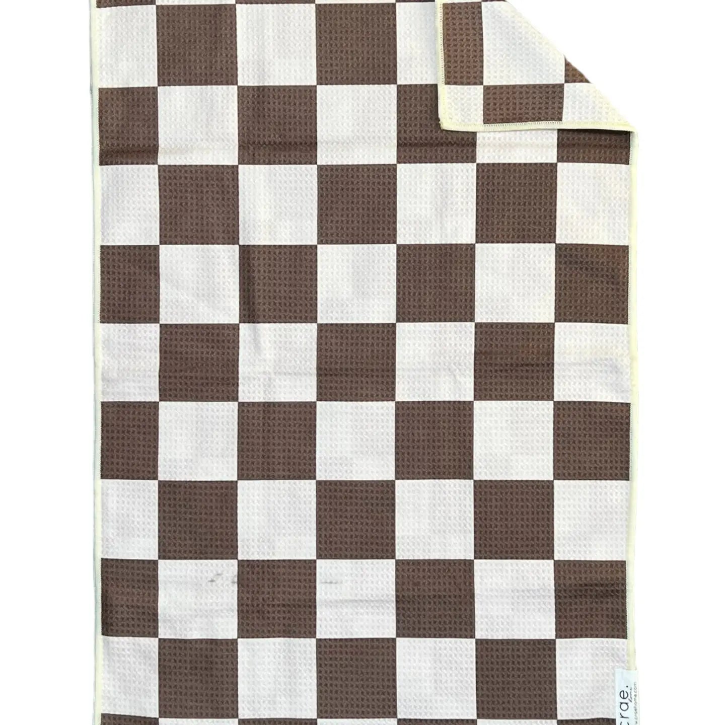 The Faire Double Sided Hand Towel features a brown and white checkerboard waffle weave design. One corner is folded to show the reverse side, with a visible small tag in the bottom right.