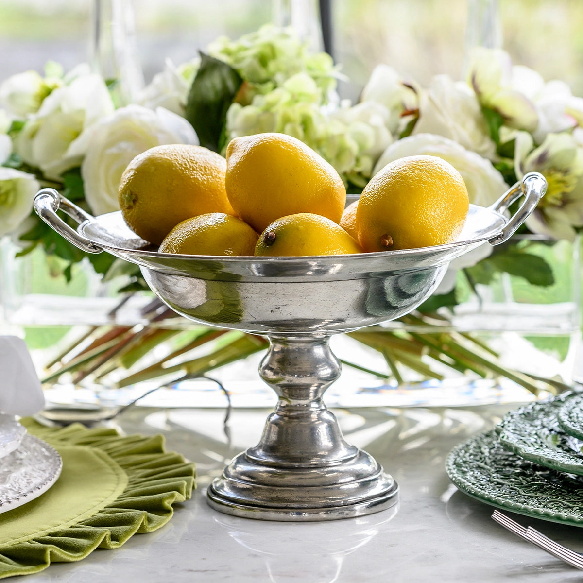 The Faire Vintage 1795 Pedestal Bowl, handmade in Italy, displays vibrant yellow lemons on a marble table accented with green plates, a matching napkin, and white flowers in the background.