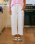 A person stands in a kitchen with hands in their pockets, wearing Frank & Eileen's Penny Sweatpant. Only their lower half is visible; they're also sporting white sandals and a pink-and-white striped top.
