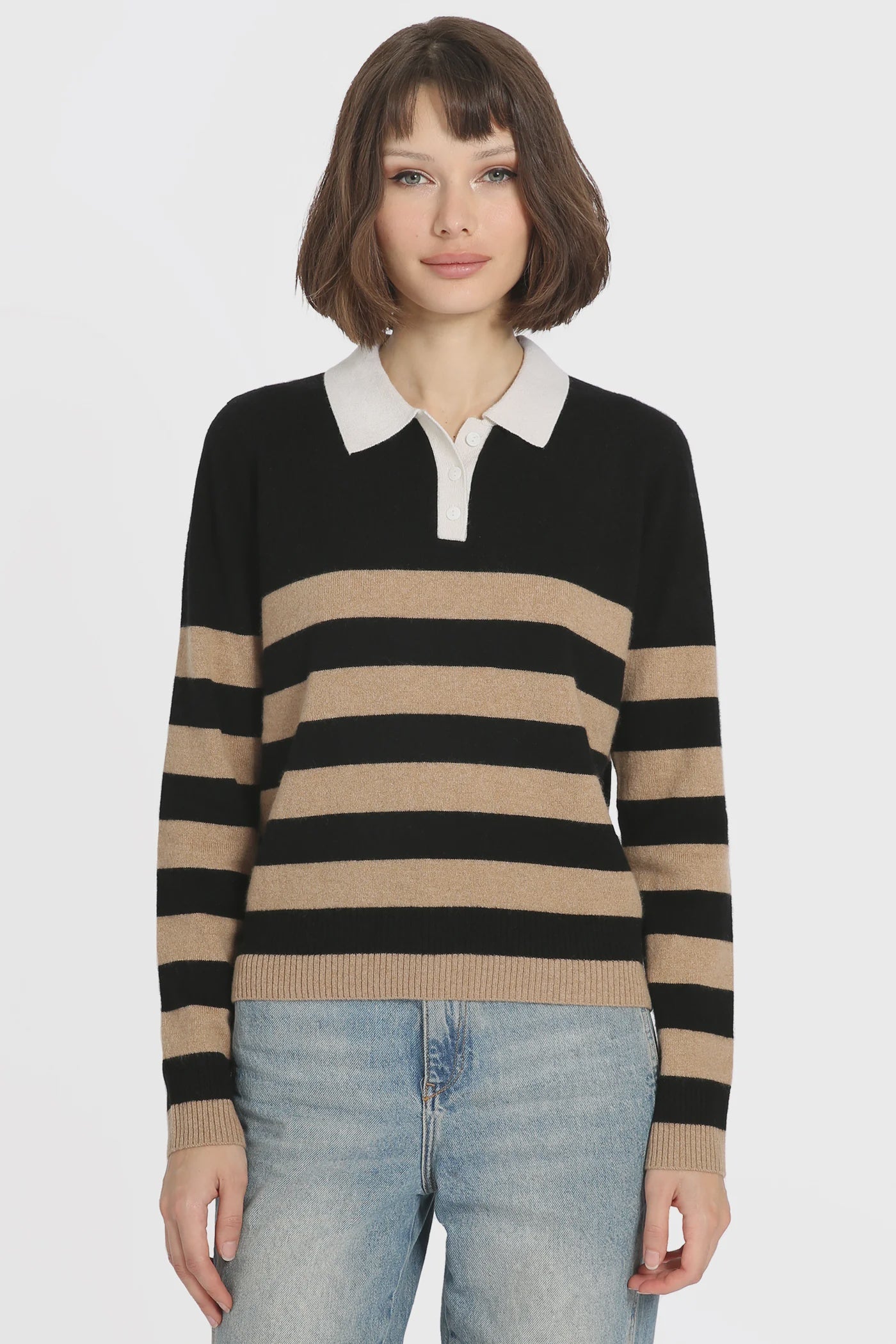 A person with short brown hair stands against a plain white background while wearing the Rugby Stripe Polo by Minnie Rose, which features classic black and tan stripes with a white collar, paired with light blue jeans.