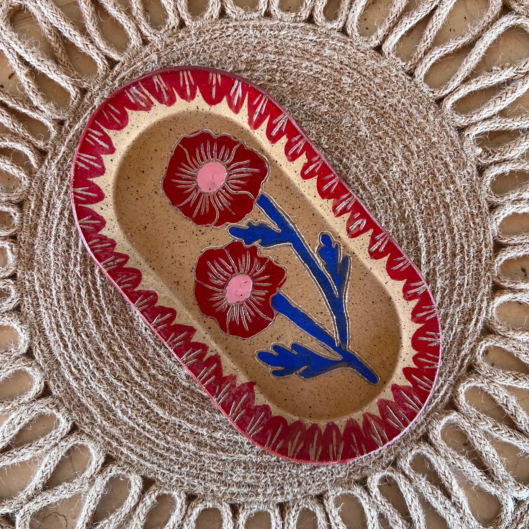 The Faire Blossom Sgraffito Dish, an oval ceramic piece with two red flowers and green stems bordered by a red pattern, is displayed on a woven sunburst placemat.