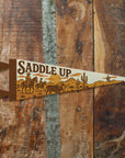 The "Saddle Up" Felt Pennant by Faire features "SADDLE UP" with cowboys, cattle, cacti, and desert scenes—a great western decor piece or horse lovers gift—shown on a rustic wooden surface.