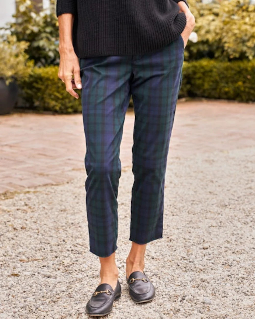 A person stands outdoors on a gravel path, wearing Frank & Eileen’s WICKLOW The Italian Chino in Italian Performance Twill, paired with a black sweater and loafers. Only their lower half is visible, with shrubs and plants in the background.