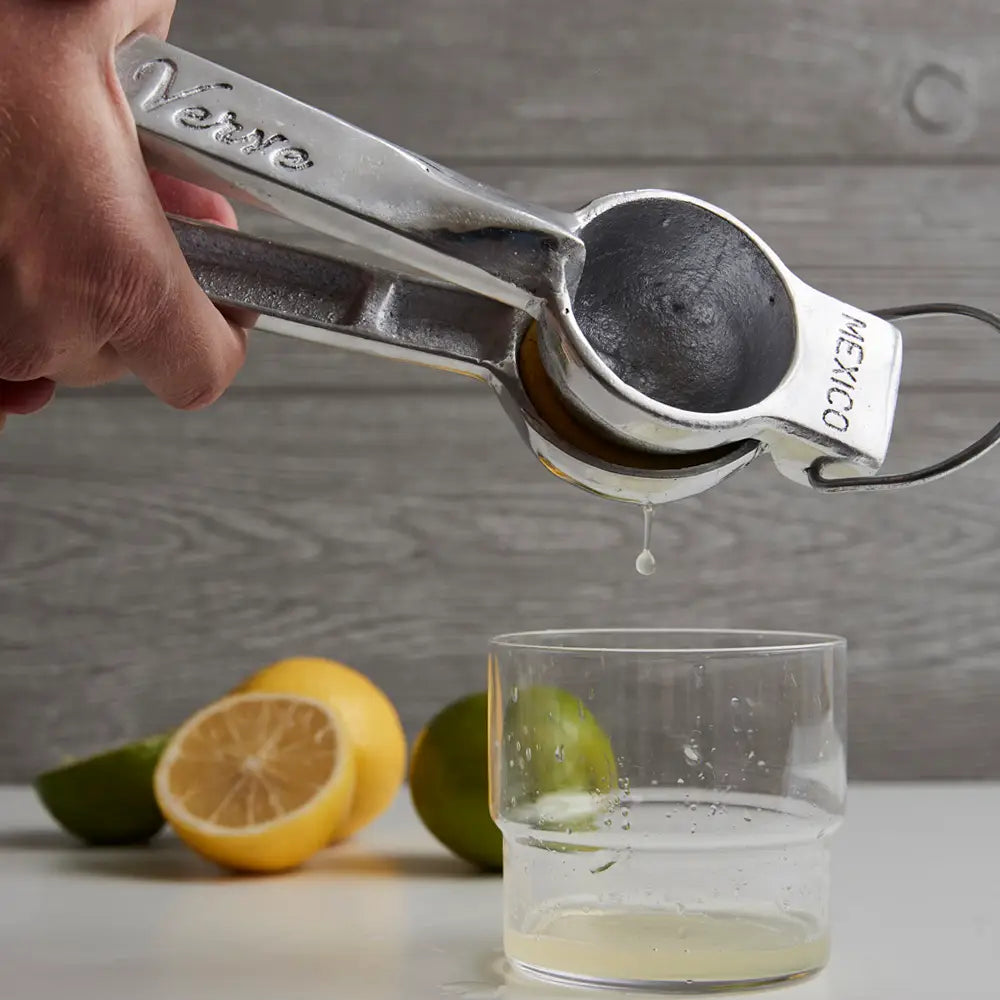 A hand squeezes lemon juice using the Faire Aluminum Hand Juicer into a clear glass, with lime and lemon halves scattered on the gray wooden surface in the background.