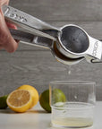 A hand squeezes lemon juice using the Faire Aluminum Hand Juicer into a clear glass, with lime and lemon halves scattered on the gray wooden surface in the background.