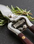 The Faire Thai Kitchen Garden Shears, featuring wooden handles and stainless steel blades, rest on a dark surface as they trim fresh rosemary.