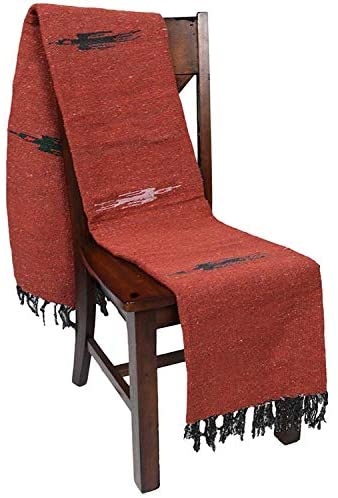 A wooden chair draped with the Faire Rust Red Baja Thunderbird Mexican Blanket. Handmade in Mexico, it features black fringe at the ends and subtle black and white patterns.