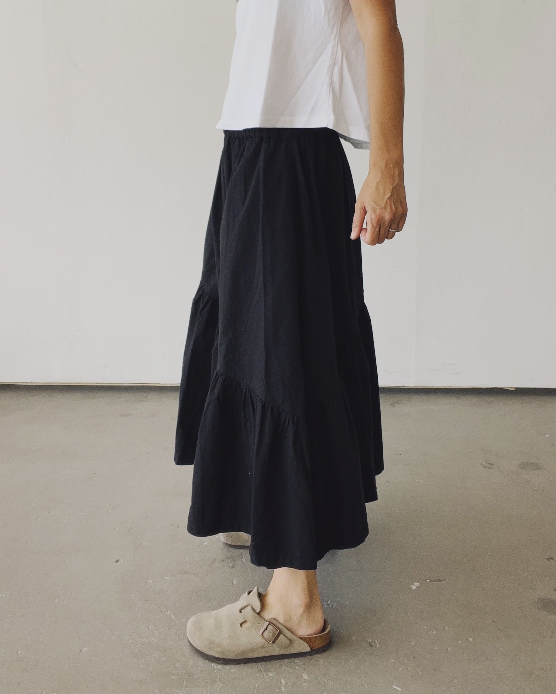 A person stands indoors in a white shirt, beige slip-on shoes, and The Ranch Skirt by Hey Gang—a long black elastic waist skirt. The side profile is shown from shoulder to feet against a plain wall.