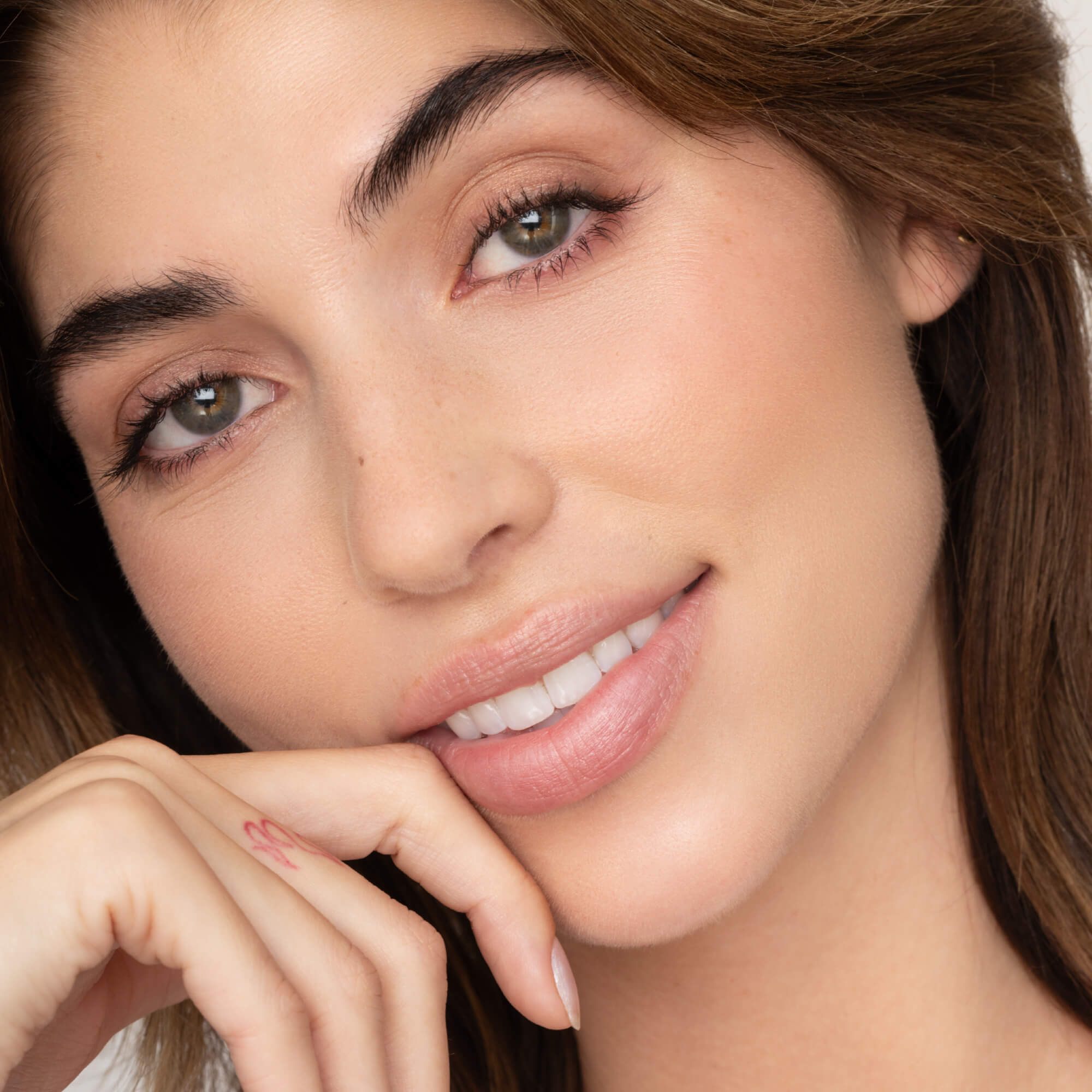 Close-up of a woman with light skin, green eyes, and brown hair, smiling gently with her chin on her hand. She showcases a small heart tattoo and soft makeup featuring Faire’s Poppy & Pout Lip Tint in the shade Farrah.