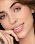 Close-up of a woman with light skin, green eyes, and brown hair, smiling gently with her chin on her hand. She showcases a small heart tattoo and soft makeup featuring Faire’s Poppy & Pout Lip Tint in the shade Farrah.
