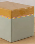 The Be Home Harlow Bright Recipe Box features a rectangular ceramic design with a glossy mustard yellow lid, pastel pink rim, and matte light gray base—ideal for organizing recipes in your kitchen and stylishly displayed on wood.