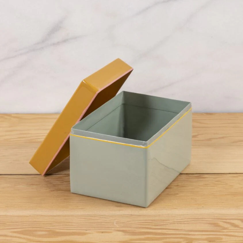 The Harlow Bright Recipe Box by Be Home, a rectangular light gray enamel box with a partially open mustard yellow lid, sits on a wooden surface and brings gourmet kitchen charm to the light marble backdrop.