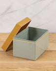 The Harlow Bright Recipe Box by Be Home, a rectangular light gray enamel box with a partially open mustard yellow lid, sits on a wooden surface and brings gourmet kitchen charm to the light marble backdrop.